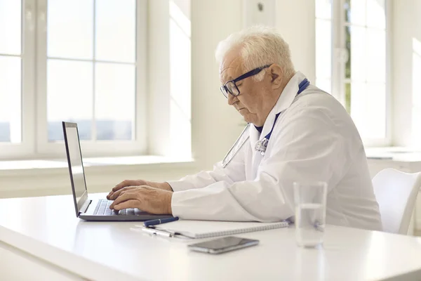 Verouderde professionele arts in uniform zitten aan de laptop en communiceren met de patiënt online — Stockfoto