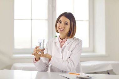 Gülümseyen tıp uzmanı elinde bir bardak su tutuyor ve susuz kalmamanı istiyor.