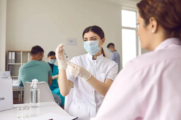 Krankenschwester in Maske wählt den Impfstoff in eine Spritze, während sie im Krankenhaus neben einem Patienten sitzt. — Stockfoto