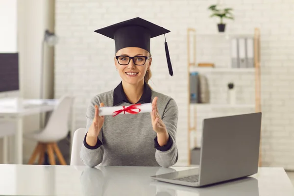 Glad online kurser examen sitter vid skrivbordet med bärbar dator, hålla hennes diplom och leende — Stockfoto