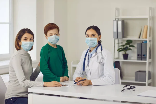 Madre, bambino e medico di famiglia in maschere viso guardando la fotocamera in ufficio ospedaliero — Foto Stock