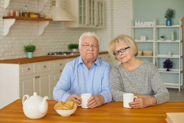 Couple âgé assis dans la cuisine regardant la caméra et boire du thé ou du café avec des cookies. — Photo