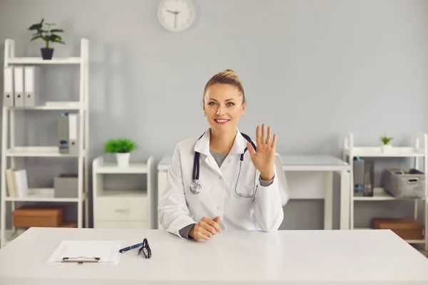 Genç, gülümseyen sarışın kadın doktor, hemşire terapist oturuyor ve hastayı elleriyle selamlıyor. — Stok fotoğraf