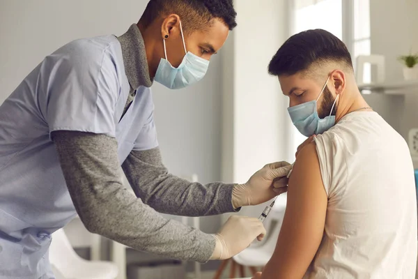 Médico afroamericano en mascarilla médica que da vacuna antiviral a un paciente joven — Foto de Stock