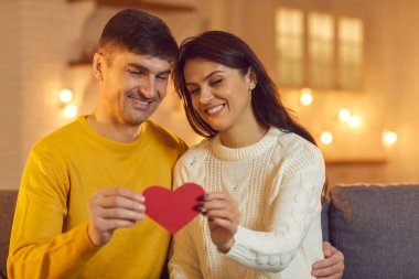 Genç, sevgi dolu mutlu çift kanepede oturuyor ve kırmızı dekoratif kalbi bir arada tutuyorlar.