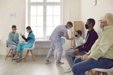 Tıbbi maskeli çeşitli insanlar sırada oturup grip ya da Covid-19 aşısı olmayı bekliyorlar.