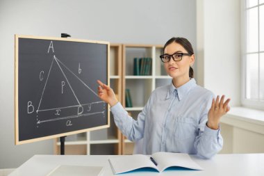 Öğretmen masasında oturuyor, tahtaya bakıyor ve öğrencilere geometri problemini nasıl çözeceklerini öğretiyor.