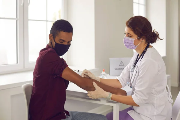 Vrouwelijke arts die vaccinatie met injectiespuit tegen coronavirusinfectie maakt voor jonge zwarte man — Stockfoto