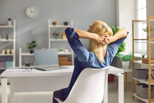 Happy senior žena relaxuje v křesle, sedí za stolem s notebookem v útulné domácí kanceláři — Stock fotografie