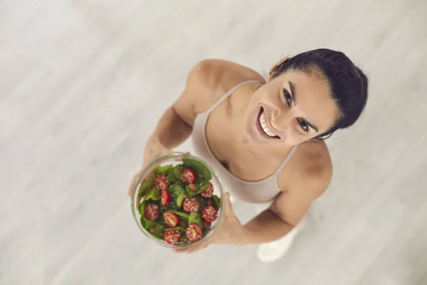 Ovanifrån av fitness kvinna innehav skål med grön basilika och körsbär tomat tittar upp på kameran. — Stockfoto