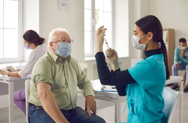 Oudere patiënt wachtend op een injectie en kijkend naar een jonge arts die het vaccin klaarmaakt — Stockfoto