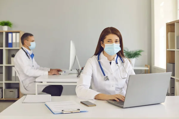 Jonge arts in medisch gezichtsmasker werkt op laptop in ziekenhuiskantoor samen met collega — Stockfoto