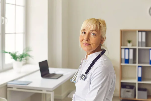 Ritratto ravvicinato di anziana stanca dottoressa che guarda la macchina fotografica mentre è in piedi nello studio medico. — Foto Stock