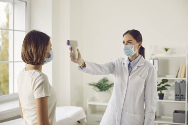 Koruyucu maskeli kadın doktor vücut ısısını elektronik termometreyle ölçüyor.