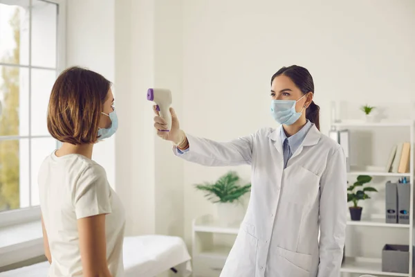 Női orvos védő maszkban méri a testhőmérsékletet elektronikus hőmérővel női beteg — Stock Fotó