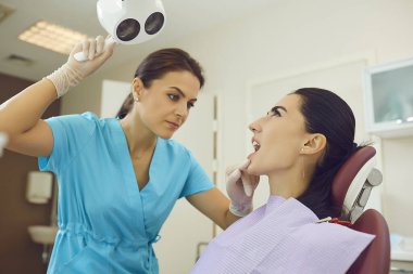 Tıbbi üniformalı dişçi muayene için kadın hastalara ışık tutuyor.
