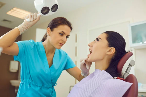 Dentist in medical uniform directing light to woman patients mouth for examination — Stock Photo, Image