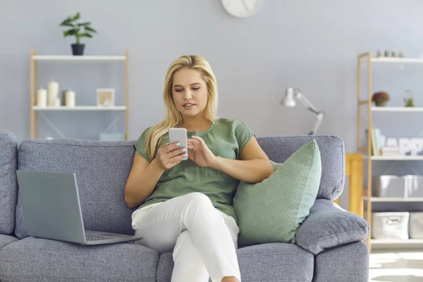 Evde dizüstü bilgisayarıyla kanepede oturan ve cep telefonu uygulaması kullanan genç bir kadın.