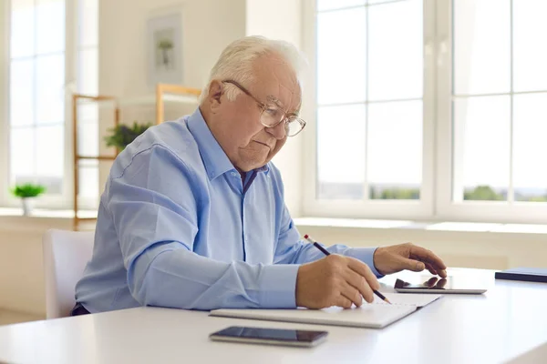Ältere Mann Arbeiter sitzt mit verschiedenen Gadgets am Schreibtisch und macht Notizen mit Bleistift bei der Arbeit — Stockfoto