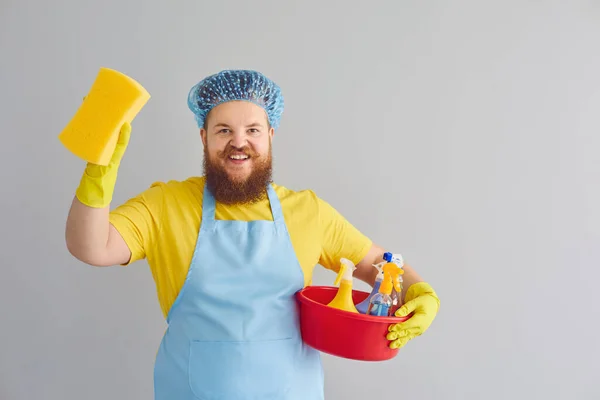 Hombre gordo divertido con barba en un delantal lavado limpia sobre un fondo gris. — Foto de Stock