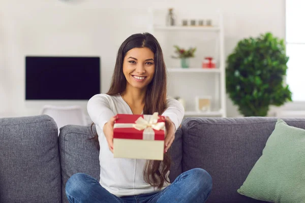 Woman sits on the couch and holds a wrapped gift in front of her and hands it to the camera. — Stock Photo, Image