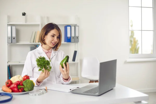 Young woman doctor nutritiologist showing fresh ingredients to patient online on laptop