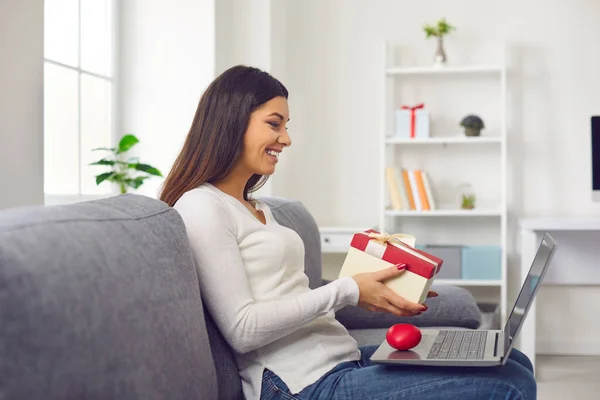 Smiling woman sitting on sofa and showing holiday present box to boyfriend during online videocall — Stock Photo, Image