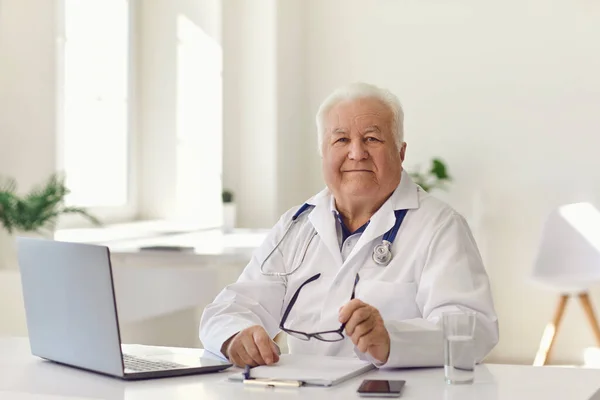 Przyjazny starszy lekarz siedzący przy biurku z laptopem w nowoczesnym gabinecie szpitalnym i patrzący w kamerę — Zdjęcie stockowe