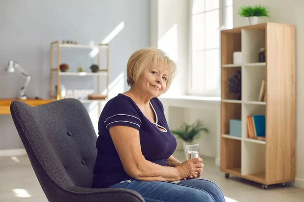 Femme retraitée positive grand-mère en jeans et t-shirt assis avec un verre d'eau dans le fauteuil — Photo