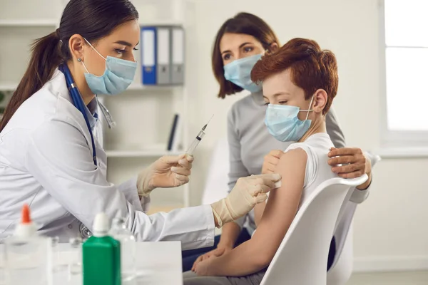 Yüz maskeli doktor annesiyle hastaneye gelen küçük çocuğa grip ya da Covid-19 aşısı yapıyor. — Stok fotoğraf