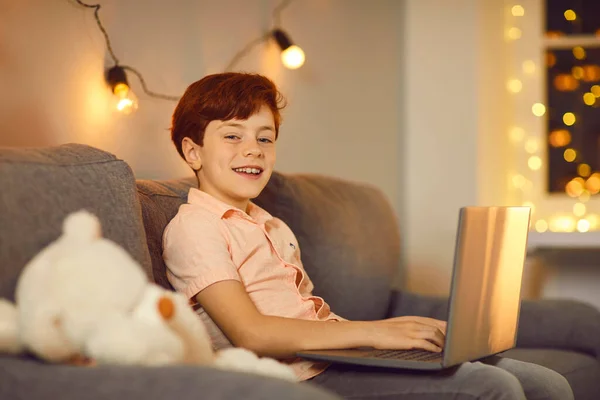 Roztomilý usměvavý chlapec sedí s notebookem doma na pohovce a korespondenci s přáteli. — Stock fotografie