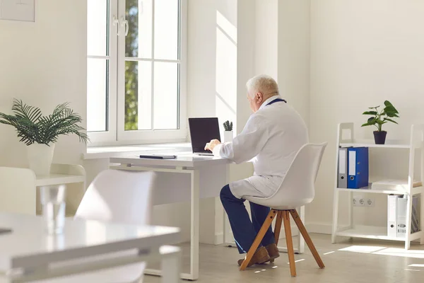 Ouderen grijs harige arts in wit uniform zitten op de laptop en communiceren met de patiënt online — Stockfoto