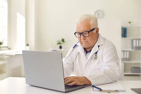 Poważny starszy lekarz piszący na klawiaturze laptopa siedzącej przy biurku w gabinecie kliniki — Zdjęcie stockowe