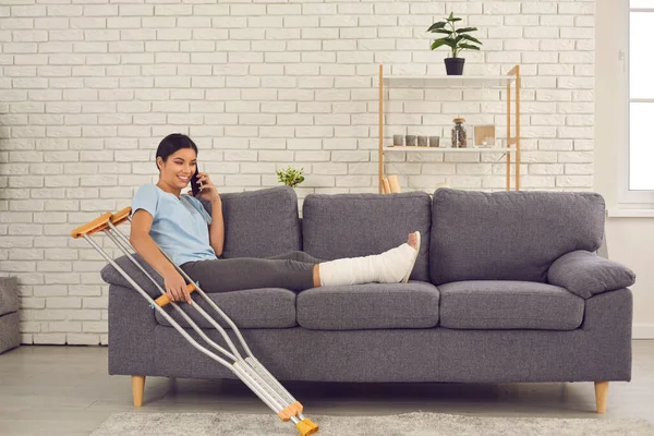Щаслива молода жінка з розбитою ногою сидить на дивані вдома і розмовляє на мобільному телефоні — стокове фото