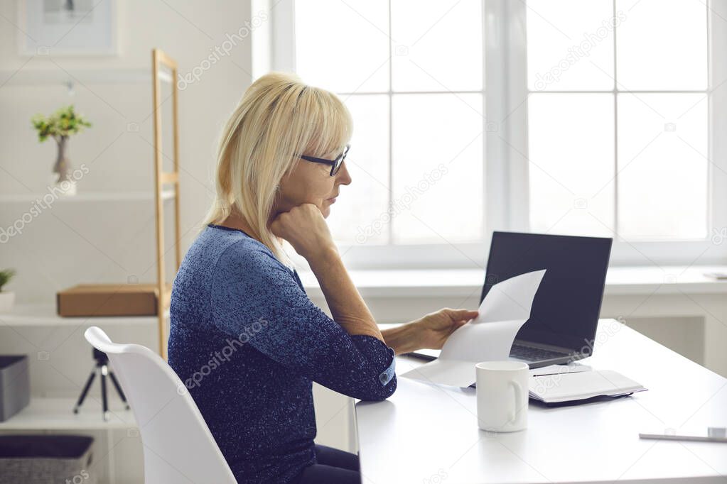Serious senior woman sitting at her desk at home and reading notification letter