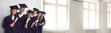 Mezunların ellerinde diplomalar ve büyük pencereli üniversite salonunda gülümseyen afişler.