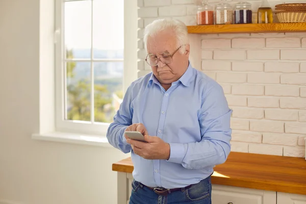 Homme âgé regarde smartphone et lit attentivement les nouvelles tout en se tenant dans la cuisine à la maison. — Photo