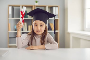 Akademik şapkalı şirin küçük kız masada oturuyor ve diplomasını elinde tutuyor.