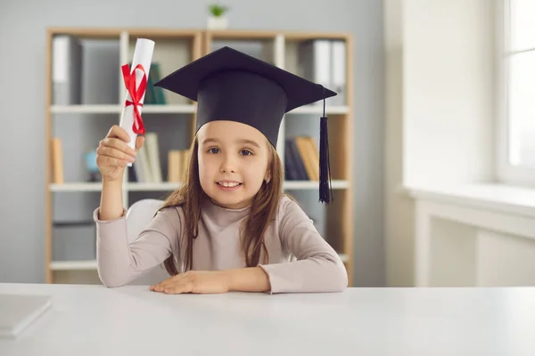 Linda niña en sombrero académico sentado en el escritorio y la celebración de su diploma de posgrado — Foto de Stock