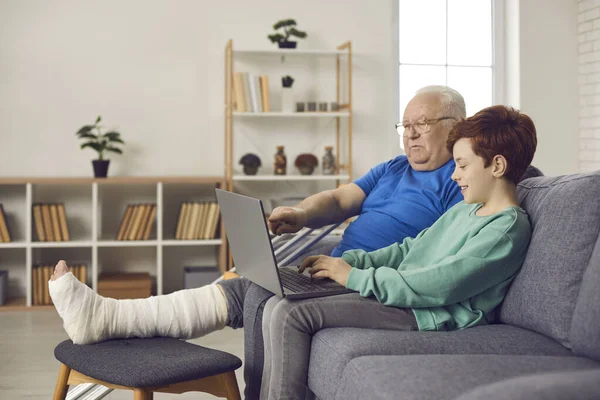 Felice nipote solidale e suo nonno con la gamba rotta utilizzando il computer portatile insieme — Foto Stock
