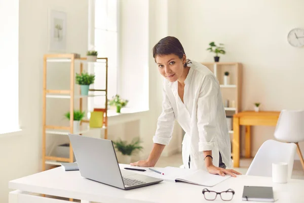 Jovem feliz inclinada na mesa com laptop aberto e planejador e olhando para a câmera — Fotografia de Stock