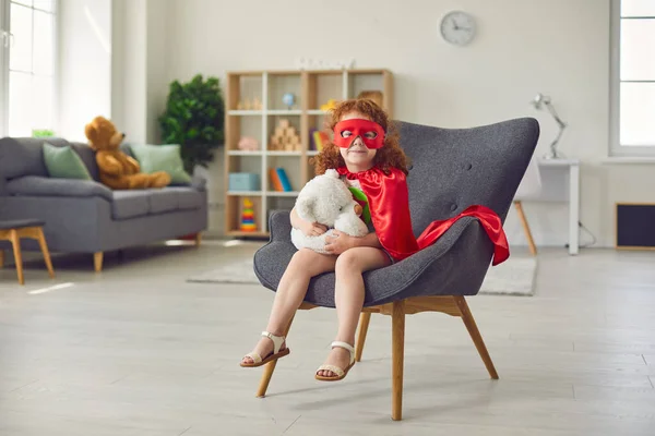 Petite fille souriante en costume de super-héros assis sur une chaise et tenant un jouet d'ours dans les mains — Photo