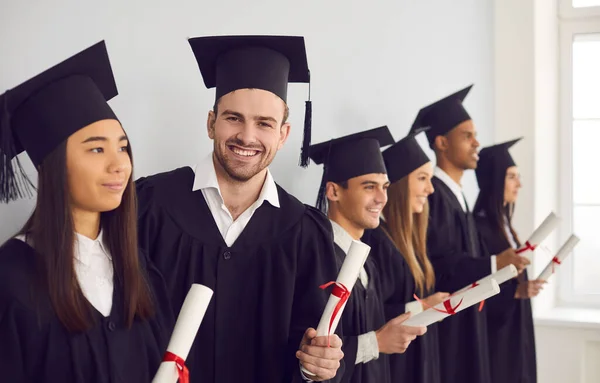 Студент с дипломом в руках смотрит в камеру, стоящую рядом со своими многонациональными одноклассниками. — стоковое фото
