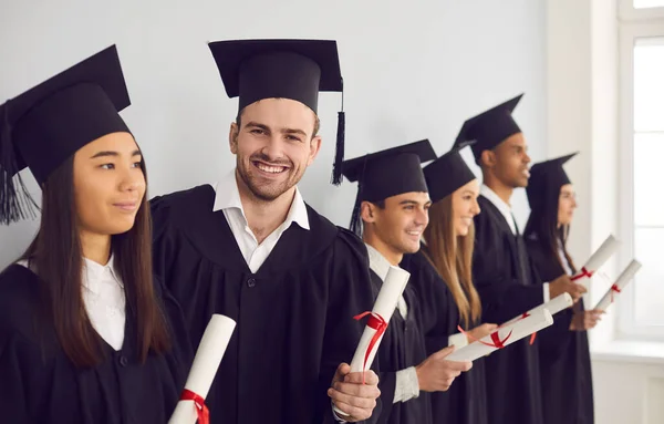 Studente uomo con diploma in mano guardando la fotocamera in piedi accanto ai suoi compagni di classe multietnici. — Foto Stock