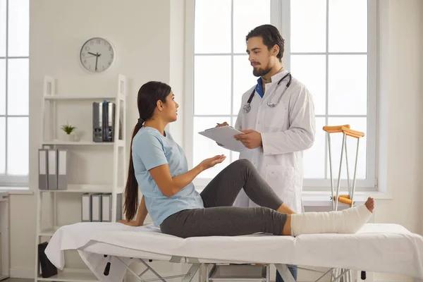 Mujer joven con la pierna rota hablando con el médico durante el examen médico en el hospital —  Fotos de Stock