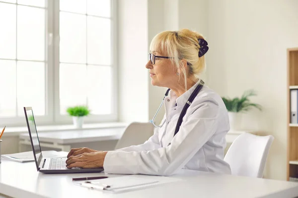 Vista lateral de una doctora sentada en su lugar de trabajo en una oficina del hospital escribiendo en una computadora portátil. — Foto de Stock