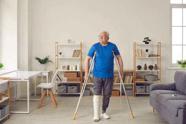 Homem sênior com a perna quebrada faz bom progresso e caminha com muletas na sala de estar — Fotografia de Stock