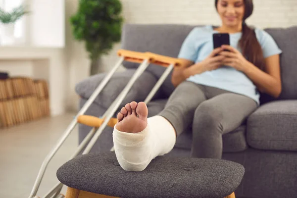 Mujer con la pierna rota en yeso yeso sentado en el sofá en casa con móvil en las manos —  Fotos de Stock