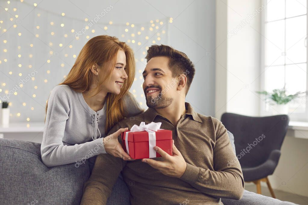 Romantic woman stands behind a man sitting on a sofa and hands him a holiday present.