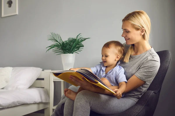 Η μητέρα διαβάζει ένα παιδικό βιβλίο στον μικρό της γιο που κάθεται σε μια καρέκλα σε ένα άνετο υπνοδωμάτιο.. — Φωτογραφία Αρχείου